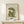 May include: Framed art print featuring olive green cut-out style figure in seated pose against cream textured background, displayed on light wood console table next to small wrapped gift with twine and greenery sprig, in minimalist neutral room setting with natural lighting and shadows.