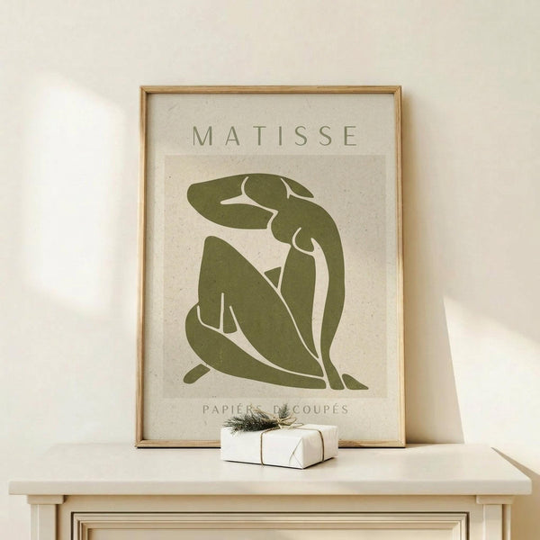 May include: Framed art print featuring olive green cut-out style figure in seated pose against cream textured background, displayed on light wood console table next to small wrapped gift with twine and greenery sprig, in minimalist neutral room setting with natural lighting and shadows.