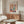 May include: Matisse-inspired art poster in thin wooden frame hanging above walnut credenza in modern living room. Poster features colorful cut-out style window scene with plants, vases, and organic shapes in coral, teal, navy, and pink. White curved chair and glass oval coffee table visible in foreground with natural lighting creating soft shadows.