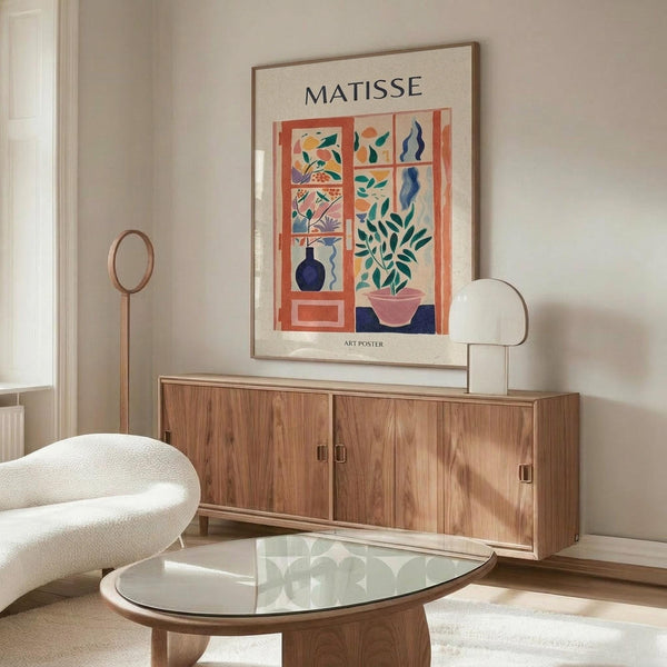 May include: Matisse-inspired art poster in thin wooden frame hanging above walnut credenza in modern living room. Poster features colorful cut-out style window scene with plants, vases, and organic shapes in coral, teal, navy, and pink. White curved chair and glass oval coffee table visible in foreground with natural lighting creating soft shadows.