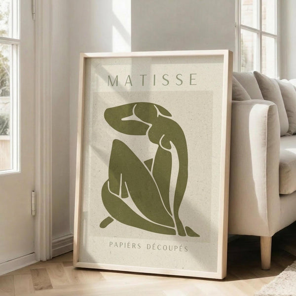 May include: Framed art print featuring olive green abstract figure silhouette in Matisse cut-out style on cream textured background, displayed in light wood frame leaning against white wall next to beige upholstered chair in bright, airy room with large windows and natural lighting casting soft shadows.