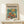 May include: Framed art print leaning against white wall on light wood shelf, featuring goldfish swimming in glass bowl with colorful flowers in teal and orange tones. Modern typography reads "matisse modernist" above the illustration. Small wrapped gift box with twine sits in front of thin natural wood frame.