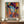 May include: Colorful portrait poster in Matisse-inspired style featuring woman wearing wide-brimmed hat, displayed in brown frame on wooden sideboard next to white ceramic vase and stacked books. Artwork shows vibrant blues, oranges, purples and geometric patterns with "MATISSE" text at bottom. Wall has white paneling in bright room setting.