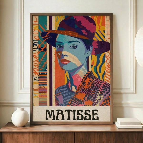 May include: Colorful portrait poster in Matisse-inspired style featuring woman wearing wide-brimmed hat, displayed in brown frame on wooden sideboard next to white ceramic vase and stacked books. Artwork shows vibrant blues, oranges, purples and geometric patterns with "MATISSE" text at bottom. Wall has white paneling in bright room setting.