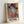 May include: Framed colorful portrait artwork leaning against white wall, featuring woman in hat rendered in vibrant blues, oranges, purples and yellows with patterned geometric elements. Light wooden frame contains vertical composition with "MATISSE" text at bottom. Bright, modern artistic style with collage-like textures and bold color blocking creates contemporary interpretation of classic portraiture.