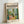May include: Framed vintage-style art poster featuring colorful modernist interior scene with bright yellow chair, green plants, orange tiled floor, and blue window frames. Light wood frame displayed against white wall corner, showing vibrant tropical garden view through windows with lush foliage and pottery.