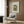 May include: Beige armchair with geometric patterned pillow, gold floor lamp, dark round side table with open book, and wood paneled wall. Framed poster shows green cut-out style figure in seated pose on cream background. Modern minimalist living room setting with natural lighting from window and warm wood tones throughout.