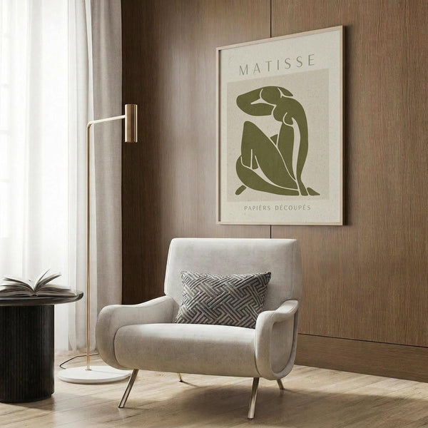 May include: Beige armchair with geometric patterned pillow, gold floor lamp, dark round side table with open book, and wood paneled wall. Framed poster shows green cut-out style figure in seated pose on cream background. Modern minimalist living room setting with natural lighting from window and warm wood tones throughout.