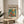 May include: Framed modernist-style poster displayed on light wood round side table next to beige upholstered chair. Artwork features colorful illustration of orange goldfish swimming among bright flowers in glass vase against teal background. Natural lighting creates soft shadows on cream walls with plant leaves visible in corner.