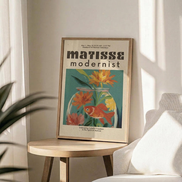 May include: Framed modernist-style poster displayed on light wood round side table next to beige upholstered chair. Artwork features colorful illustration of orange goldfish swimming among bright flowers in glass vase against teal background. Natural lighting creates soft shadows on cream walls with plant leaves visible in corner.
