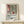 May include: Matisse-inspired poster in natural wood frame displaying colorful still life with potted plant, vases, and geometric shapes in terracotta, green, and blue tones. Text reads "Celebrating Colors of Life" with descriptive paragraph below. Frame sits on light wood surface next to small wrapped gift with twine and greenery sprig against cream wall.