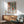 May include: Matisse-inspired poster with colorful still life featuring orange vase with leafy plant, white vase, and geometric shapes in warm terracotta, green, and cream tones. Wooden frame hangs above mid-century modern sideboard with white table lamp. Sage green upholstered chair with wooden arms sits nearby in bright, naturally lit room with white walls.