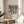 May include: Framed poster with Matisse-inspired still life artwork featuring terracotta vase with green foliage, geometric shapes in orange and blue tones, and white vase. Text reads "Celebrating Colors of Life" below colorful illustration. Wood frame sits on round wooden side table next to beige upholstered chair with plant leaves visible in corner of bright, sunlit room.