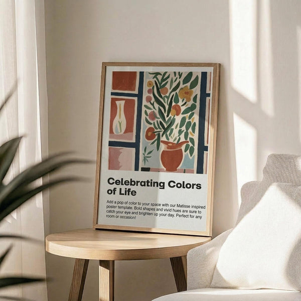 May include: Framed poster with Matisse-inspired still life artwork featuring terracotta vase with green foliage, geometric shapes in orange and blue tones, and white vase. Text reads "Celebrating Colors of Life" below colorful illustration. Wood frame sits on round wooden side table next to beige upholstered chair with plant leaves visible in corner of bright, sunlit room.