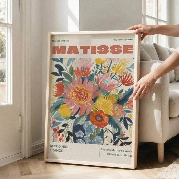May include: Hands holding a framed poster featuring vibrant floral artwork with orange, yellow, pink and blue flowers and green leaves. The poster has "MATISSE" in large red letters at the top and "06200 NICE, FRANCE" at the bottom. Light wood frame shown in bright living room with beige sofa, white walls, and large windows casting natural sunlight on hardwood floors.