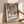 May include: Hands holding a framed poster with Matisse-inspired artwork featuring colorful vases with flowers and foliage in terracotta, green, and blue tones. The poster has "Celebrating Colors of Life" text and descriptive copy below the artwork. Natural wood frame against white wall with soft lighting and shadows.