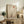May include: Framed minimalist landscape poster with beige background showing layered tan mountains, bare black tree silhouette, large white circle and smaller rust-colored circle representing celestial bodies. Light wood frame displayed on rustic wooden credenza alongside fern plant, pottery vase, and books in neutral-toned living room setting.