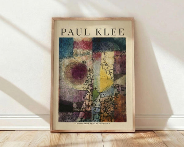 May include: Framed Paul Klee art print leaning against white wall on light wood floor. Abstract colorful artwork features geometric shapes, architectural elements, and muted tones of blue, red, yellow, purple, and green with intricate line drawings. Natural wood frame with cream matting. Soft natural lighting creates diagonal shadows across the scene.