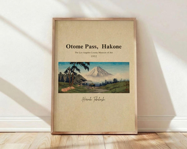 May include: Framed vintage Japanese woodblock print showing Mount Fuji in background with traditional thatched-roof house, palm trees, and rolling green hills in foreground. Artwork features soft blue sky, muted earth tones, and cream matting with museum attribution text. Natural wood frame leans against white wall on light wooden floor with soft natural lighting creating diagonal shadows.