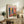 May include: Framed Matisse exhibition poster displayed on rustic wooden sideboard in cozy living room. Poster features vibrant interior scene with colorful plants, blue shutters, and bright geometric patterns in pink, yellow, orange, and green. Room setting shows cream upholstered chair, fern plant, ceramic vase, stacked books, and warm neutral walls with natural lighting creating soft shadows.