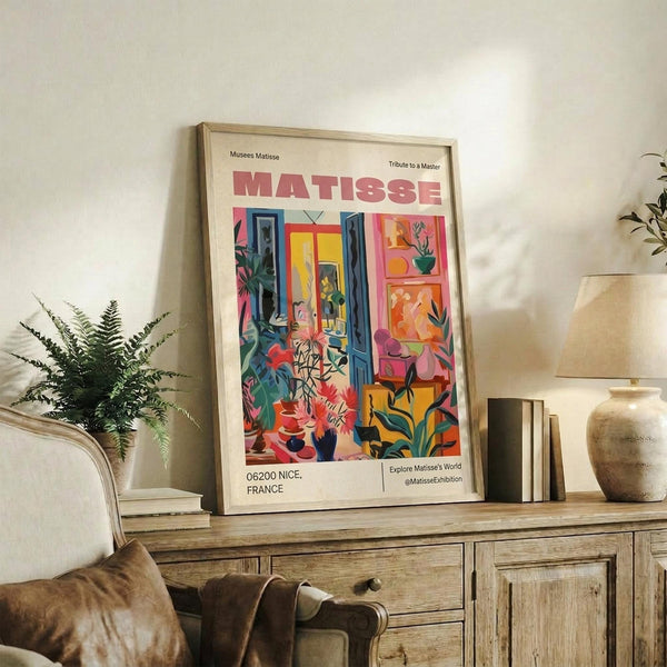 May include: Framed Matisse exhibition poster displayed on rustic wooden sideboard in cozy living room. Poster features vibrant interior scene with colorful plants, blue shutters, and bright geometric patterns in pink, yellow, orange, and green. Room setting shows cream upholstered chair, fern plant, ceramic vase, stacked books, and warm neutral walls with natural lighting creating soft shadows.