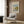 May include: Framed Claude Monet exhibition poster featuring sailboats on blue water with reflections, displayed on wood-paneled wall above cream upholstered armchair with geometric patterned pillow. Modern brass floor lamp and dark round side table with open book complete the neutral-toned interior setting with natural light from window.