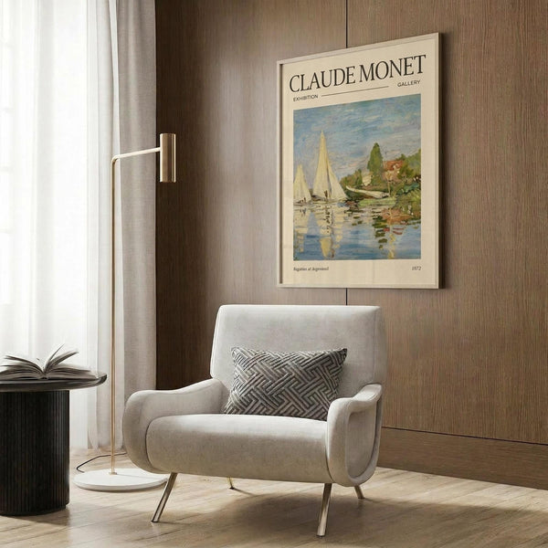 May include: Framed Claude Monet exhibition poster featuring sailboats on blue water with reflections, displayed on wood-paneled wall above cream upholstered armchair with geometric patterned pillow. Modern brass floor lamp and dark round side table with open book complete the neutral-toned interior setting with natural light from window.