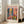 May include: Framed art print leaning against white wall next to beige sofa in bright living room. Colorful Matisse-style poster features vibrant interior scene with blue doors, pink walls, tropical plants, and flowers in bold fauve colors. Light wood frame, cream matting, hardwood floors with natural window light.