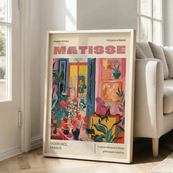 May include: Framed art print leaning against white wall next to beige sofa in bright living room. Colorful Matisse-style poster features vibrant interior scene with blue doors, pink walls, tropical plants, and flowers in bold fauve colors. Light wood frame, cream matting, hardwood floors with natural window light.