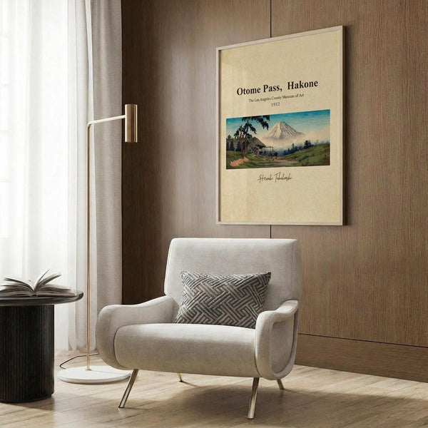 May include: Vintage Japanese travel poster print featuring Mount Fuji landscape with traditional buildings and green hills, displayed in cream-colored frame on wood paneled wall above modern beige armchair with geometric patterned pillow, brass floor lamp, and dark round side table with open book in contemporary living room setting.