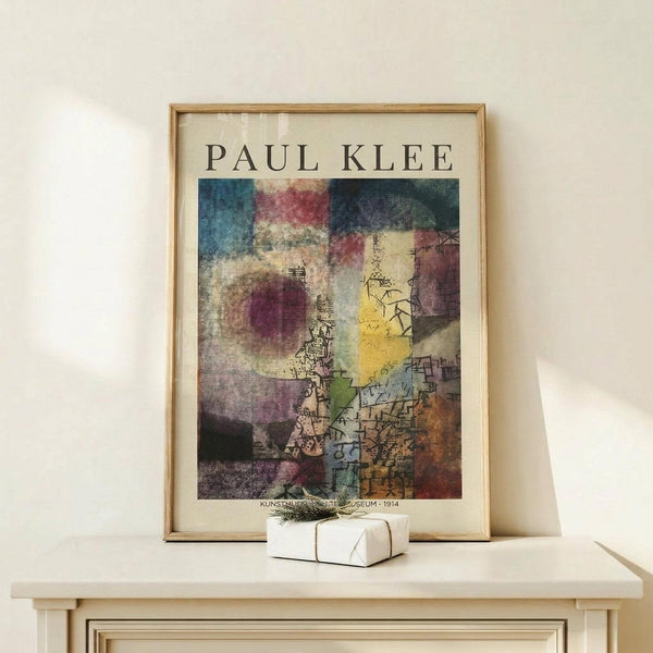 May include: Framed Paul Klee abstract art print displayed on light wood console table against cream wall. Colorful modernist artwork features geometric shapes, architectural elements, and vibrant patches of blue, red, yellow, and purple with intricate line drawings. Natural wood frame surrounds the museum poster. Small white wrapped gift with evergreen sprig sits beside the frame, creating cozy home decor scene with soft natural lighting.