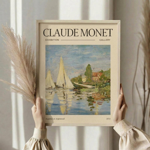 May include: Hands holding a framed Claude Monet exhibition poster featuring his 1872 painting "Regattas at Argenteuil" showing sailboats on blue water with reflections and green trees. One hand also holds dried wheat stalks. The poster has cream matting with black text and sits against white curtains in soft natural lighting.