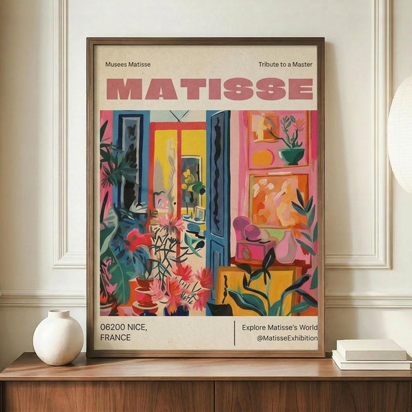 May include: Framed art print featuring vibrant Matisse-style interior scene with colorful plants, furniture, and artwork in pink, blue, yellow, and green tones. Print displays "MATISSE" text and "06200 NICE, FRANCE" at bottom. Frame sits on wooden surface beside white ceramic vase and stacked books against cream wall with decorative molding.