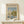 May include: Framed Claude Monet exhibition poster featuring sailboats on water with reflections, displayed on light wood surface against cream wall. Small white wrapped gift with twine and greenery sits beside the poster. Soft natural lighting creates gentle shadows on the minimalist scene.