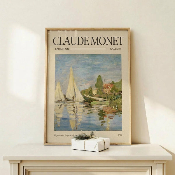 May include: Framed Claude Monet exhibition poster featuring sailboats on water with reflections, displayed on light wood surface against cream wall. Small white wrapped gift with twine and greenery sits beside the poster. Soft natural lighting creates gentle shadows on the minimalist scene.
