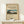 May include: Framed vintage Japanese landscape print showing Mount Fuji with rural village scene in foreground, displayed on light wood console table beside small white wrapped gift with twine and greenery sprig, against cream colored wall with natural lighting