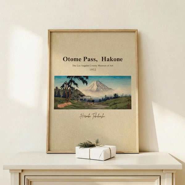 May include: Framed vintage Japanese landscape print showing Mount Fuji with rural village scene in foreground, displayed on light wood console table beside small white wrapped gift with twine and greenery sprig, against cream colored wall with natural lighting