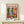 May include: Colorful Matisse exhibition poster in natural wood frame on light wooden surface. Vibrant interior scene shows blue doors opening to yellow room, tropical plants, red flowers in vases, and artwork on pink walls. Small wrapped gift with greenery sits beside frame against cream wall.