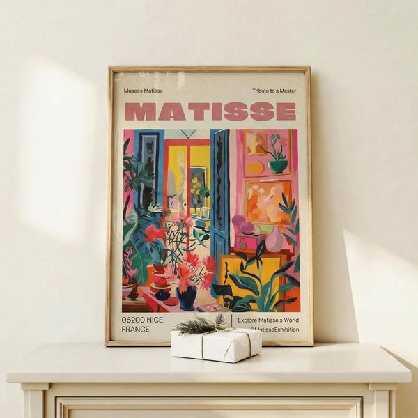 May include: Colorful Matisse exhibition poster in natural wood frame on light wooden surface. Vibrant interior scene shows blue doors opening to yellow room, tropical plants, red flowers in vases, and artwork on pink walls. Small wrapped gift with greenery sits beside frame against cream wall.