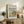 May include: Framed Claude Monet exhibition poster featuring sailboats on water with reflections, displayed on rustic wood sideboard alongside fern plant, books, and ceramic vases in neutral-toned living room setting with beige upholstered chair visible.