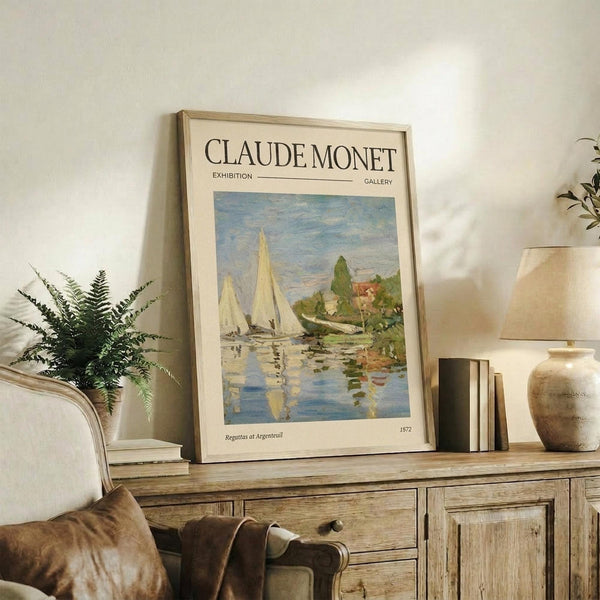 May include: Framed Claude Monet exhibition poster featuring sailboats on water with reflections, displayed on rustic wood sideboard alongside fern plant, books, and ceramic vases in neutral-toned living room setting with beige upholstered chair visible.