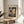 May include: Framed Paul Klee art print displayed on round wooden side table next to beige upholstered chair. Abstract colorful artwork features geometric shapes in blues, reds, yellows, and earth tones with linear drawings overlaid. Natural sunlight creates shadows across neutral wall. Green plant leaves visible in corner of minimalist room setting.
