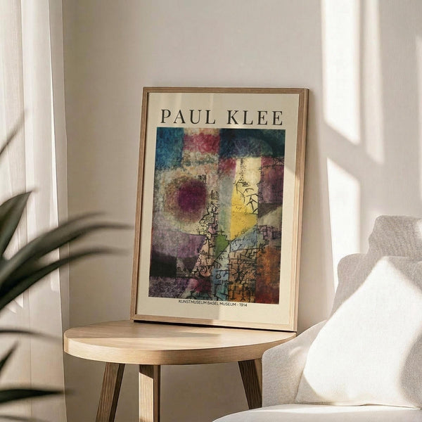 May include: Framed Paul Klee art print displayed on round wooden side table next to beige upholstered chair. Abstract colorful artwork features geometric shapes in blues, reds, yellows, and earth tones with linear drawings overlaid. Natural sunlight creates shadows across neutral wall. Green plant leaves visible in corner of minimalist room setting.