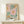 May include: Framed botanical art print featuring stylized flowers in pink, orange, and yellow with green foliage on cream background, displaying "NO RAIN NO FLOWERS" text at bottom. Light wood frame sits on pale wooden surface beside small wrapped gift with twine and greenery sprig against neutral wall.