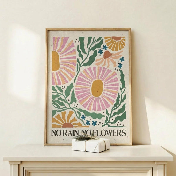 May include: Framed botanical art print featuring stylized flowers in pink, orange, and yellow with green foliage on cream background, displaying "NO RAIN NO FLOWERS" text at bottom. Light wood frame sits on pale wooden surface beside small wrapped gift with twine and greenery sprig against neutral wall.