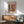 May include: Colorful Matisse exhibition poster in wooden frame hanging above mid-century modern credenza with white ceramic lamp. Poster features vibrant interior scene with blues, reds, yellows, and greens depicting a studio space with plants and furniture. Green upholstered armchair with wooden frame sits nearby on light wooden floor with natural sunlight casting shadows across white wall.