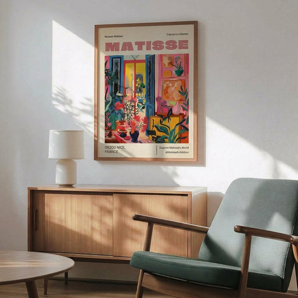 May include: Colorful Matisse exhibition poster in wooden frame hanging above mid-century modern credenza with white ceramic lamp. Poster features vibrant interior scene with blues, reds, yellows, and greens depicting a studio space with plants and furniture. Green upholstered armchair with wooden frame sits nearby on light wooden floor with natural sunlight casting shadows across white wall.