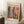 May include: Framed vintage-style art print featuring Henri Matisse exhibition poster with colorful interior scene showing blue doors, tropical plants, vases, and artwork. Print displays "MATISSE" in large pink letters, "06200 NICE, FRANCE" and "Musees Matisse" text. Light wood frame leans against white wall beside coffee cup on wooden block and textured throw blanket on cream rug.