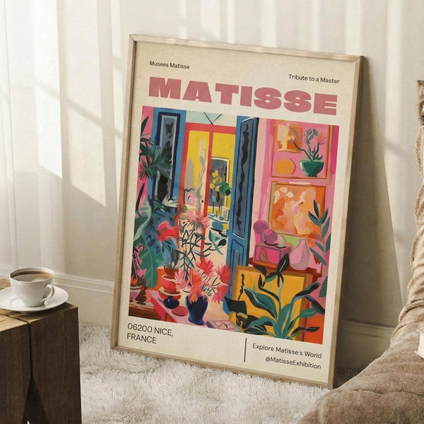 May include: Framed vintage-style art print featuring Henri Matisse exhibition poster with colorful interior scene showing blue doors, tropical plants, vases, and artwork. Print displays "MATISSE" in large pink letters, "06200 NICE, FRANCE" and "Musees Matisse" text. Light wood frame leans against white wall beside coffee cup on wooden block and textured throw blanket on cream rug.