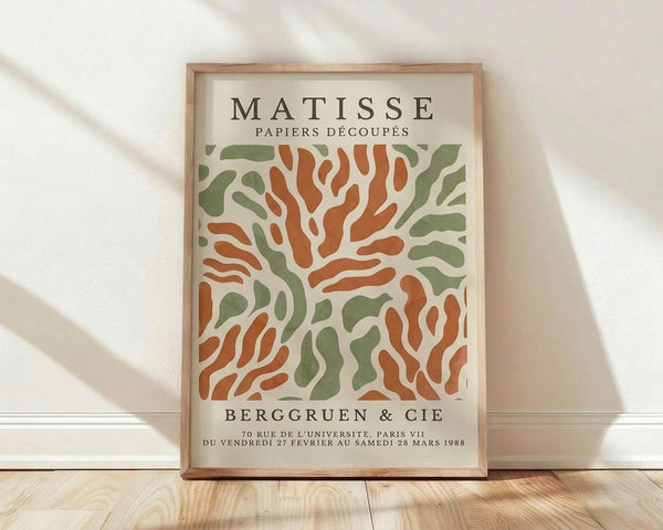 May include: Framed vintage exhibition poster featuring Henri Matisse's cut-out style artwork in organic coral orange and sage green shapes on cream background, displayed against white textured wall on light wood floor with natural shadow patterns